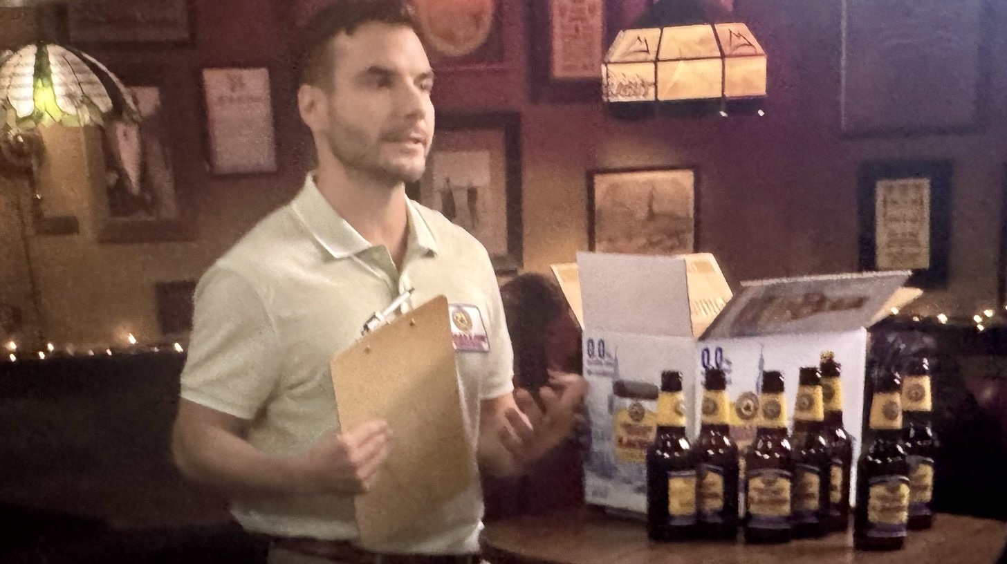 A beer sales rep with a clipboard stands in a dim bar beside an open case of nonalcoholic beer bottles.